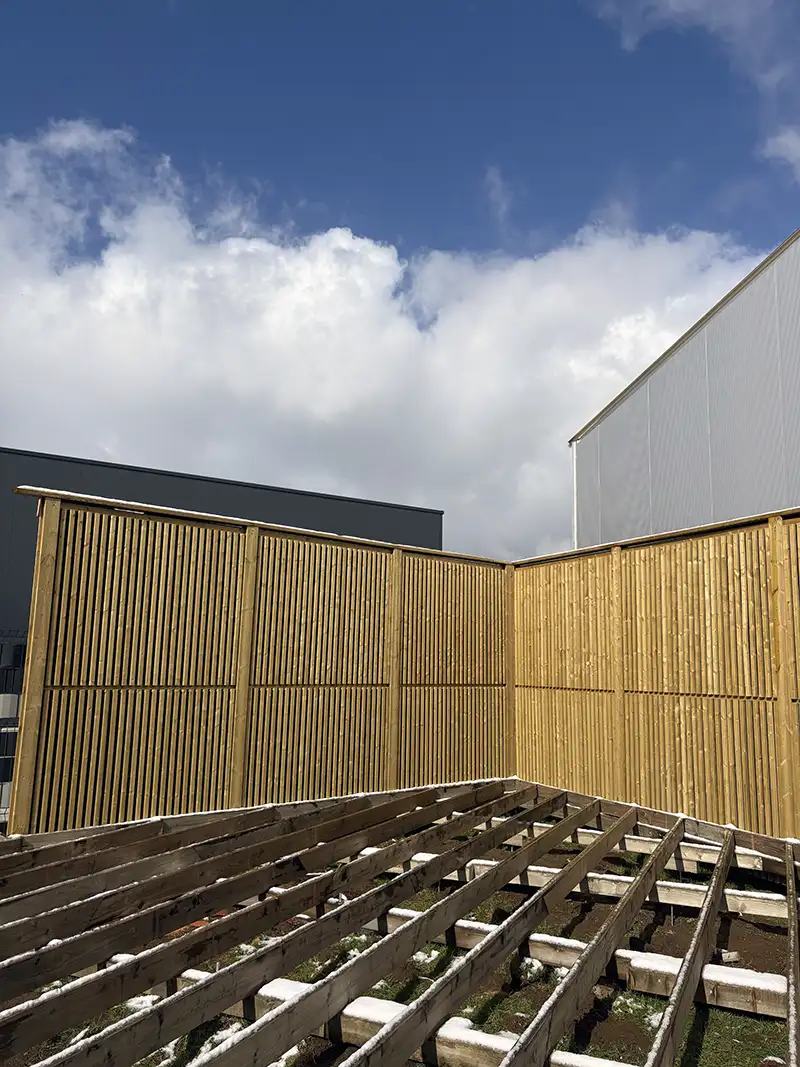Des panneaux de bois clairs sont disposés verticalement, encadrant une structure au sol sous un ciel bleu avec quelques nuages.