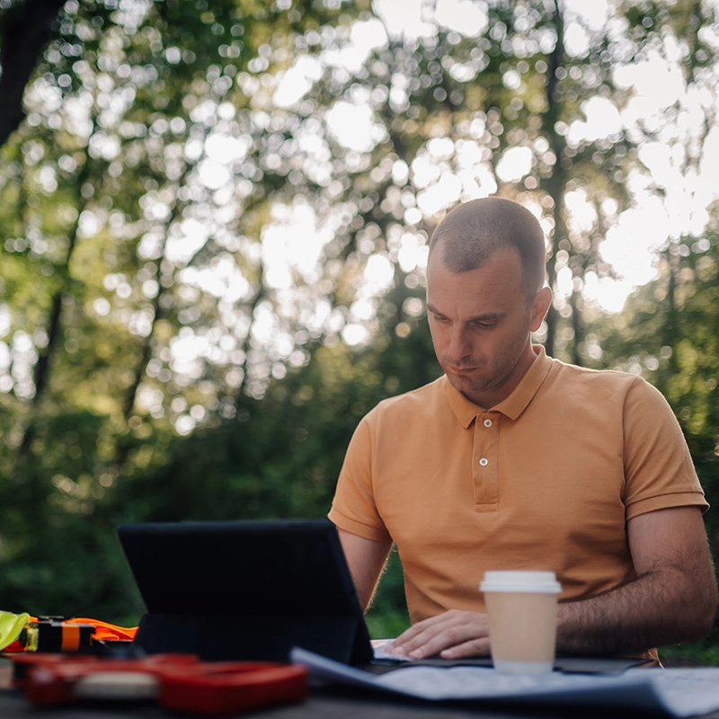 Un homme en polo orange utilise une tablette à l'extérieur, avec des arbres en arrière-plan et un gobelet en carton sur la table.