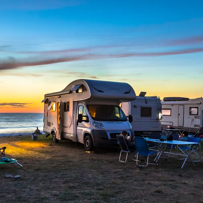 Camping-car blanc stationné près de la mer au coucher du soleil, avec des chaises et une table de camping à côté.