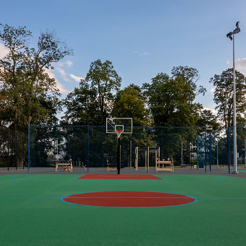 Terrain de basketball extérieur avec sol vert et rouge, entouré d'arbres et éclairé par un lampadaire.