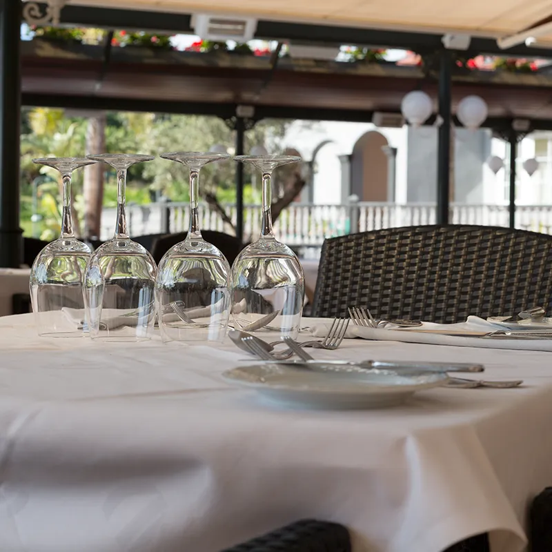 Quatre verres à pied retournés sur une table dressée avec une nappe blanche et des couverts, en terrasse.