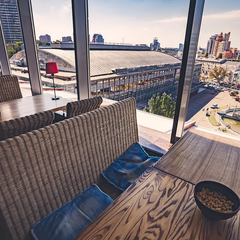 Table en bois avec chaises en rotin, lampe rouge, vue sur des bâtiments urbains à travers de grandes fenêtres vitrées.