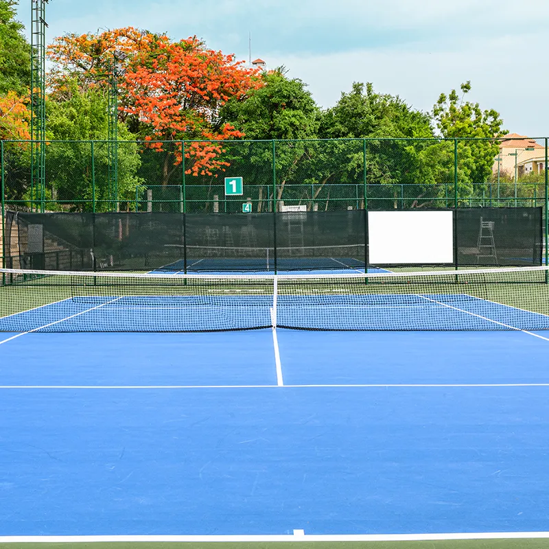Terrain de tennis bleu avec filet au centre, entouré d'arbres verts et orange, sous un ciel dégagé.