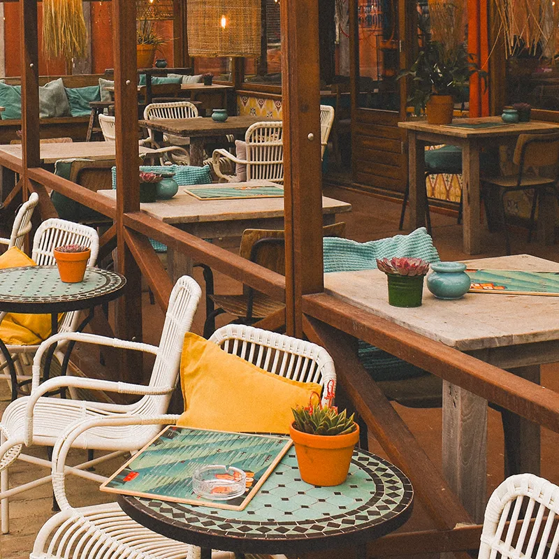 Terrasse de restaurant avec tables en bois, chaises blanches, coussins jaunes, et plantes en pot sur les tables.
