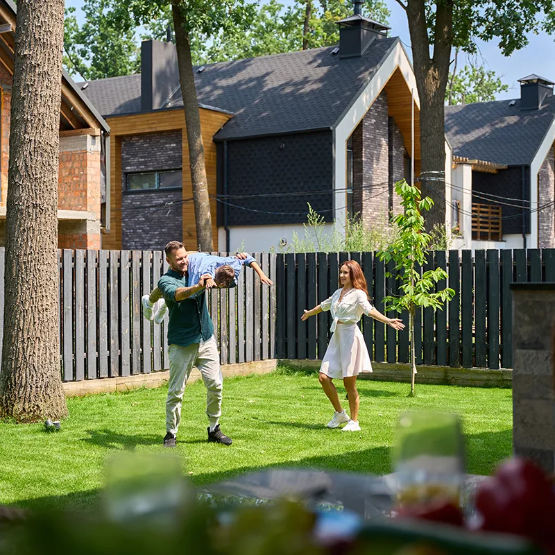 Un couple joue avec un enfant dans un jardin, entouré de maisons modernes et d'arbres, près d'une clôture en bois.