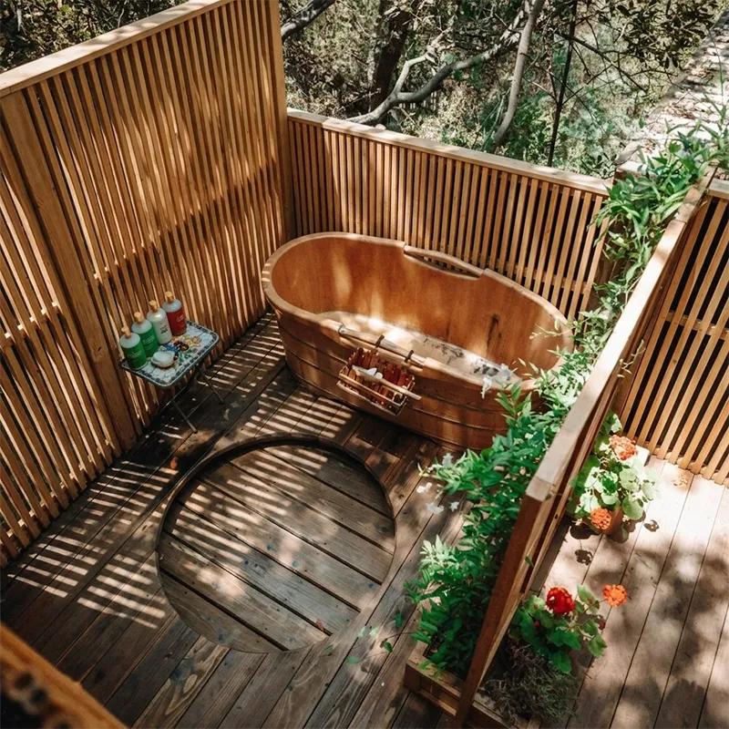 Baignoire en bois entourée de cloisons en bois, avec des plantes vertes et des fleurs rouges à côté.