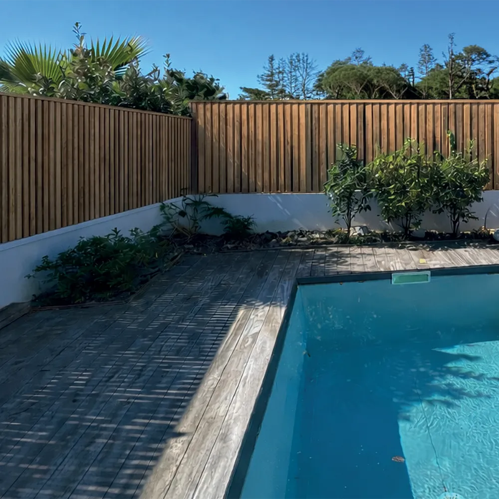 gamme-privilege Un jardin avec pelouse verte, chemin de dalles, clôture en bois, et terrasse aménagée avec chaises longues près d'une maison.